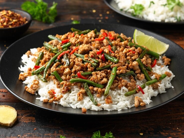 Ein schwarzer Teller mit der asiatischen Hackpfanne auf Reis. Alles auf einem dunkelbraunen Tisch mit Ingwer und Limette.