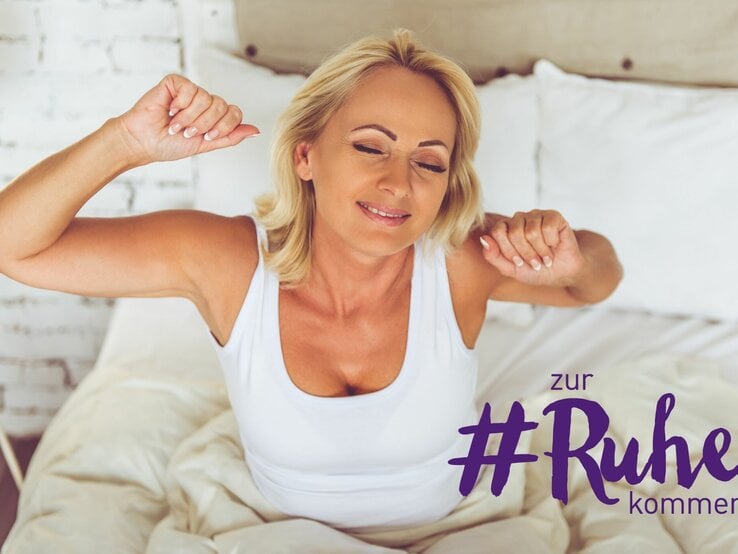 Eine blonde Frau in einem weißen Tanktop dehnt sich gerade nach dem Aufwachen genüsslich im Bett. Sie hat die Augen geschlossen und ein entspanntes Lächeln auf den Lippen, während sie ihre Arme nach oben und hinten streckt. Das Bettzeug ist neutral und die Umgebung wirkt beruhigend mit einer hellen und luftigen Atmosphäre. Der Schriftzug "+#zur Ruhe kommen" in lila Farbe ist in der Ecke des Bildes platziert und unterstreicht das Thema der Entspannung und des Wohlbefindens.