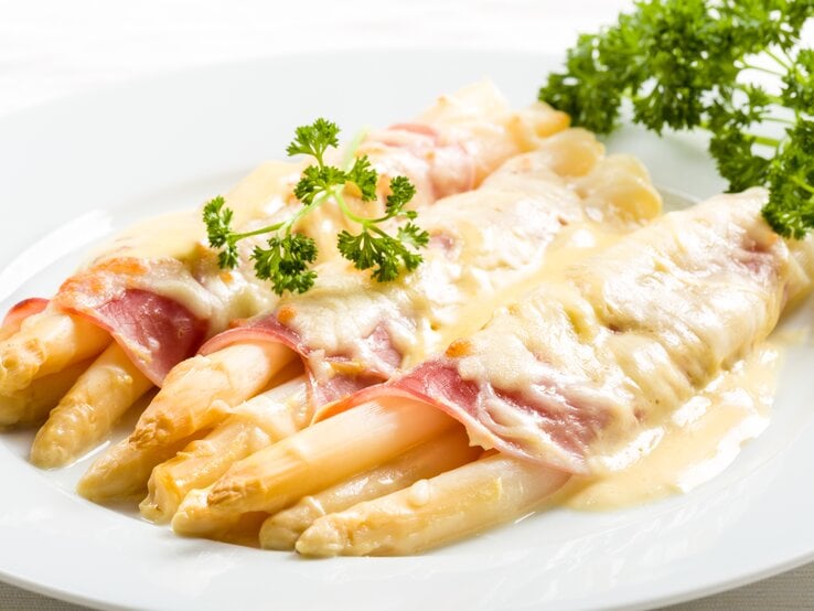 Weißer Spargel überbacken mit Schinken und Sauce hollandaise. Daneben liegt frische Petersilie.