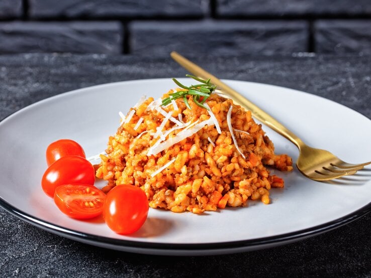 Hackfleisch-Risotto mit Cocktailtomaten auf weißem Teller mit Kupfergabel auf dunklem Untergrund. Draufsicht.