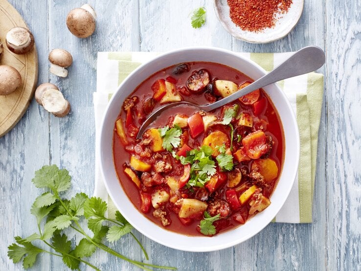 Gemüseeintopf mit Hack und Chili, bestehend aus Tomatenbrühe, Knollengemüse, kross gebratenem Hackfleisch und einer Garnitur aus Petersilie und Kroiander. Alles serviert in einer großen Suppenschale und garniert mit Chiliflocken. 