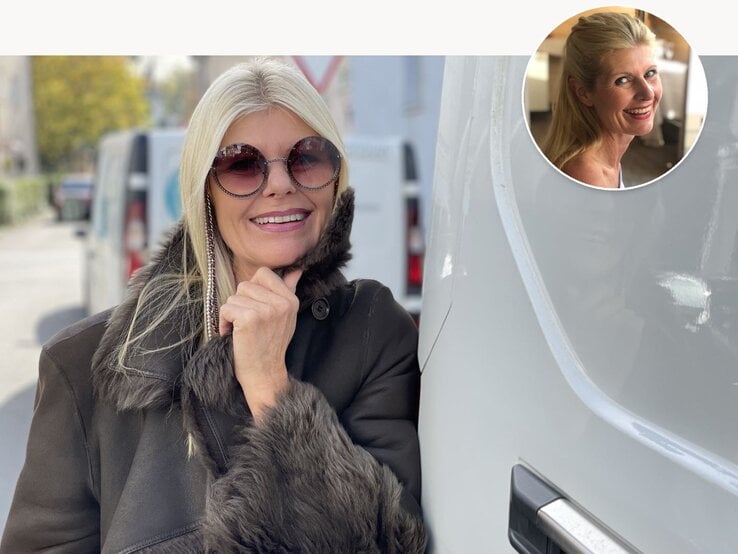 Frau mit langem blondem Haar und schwarzem Mantel mit Fellkragen lehnt nachdenklich an weißem Fahrzeug auf sonniger Straße.