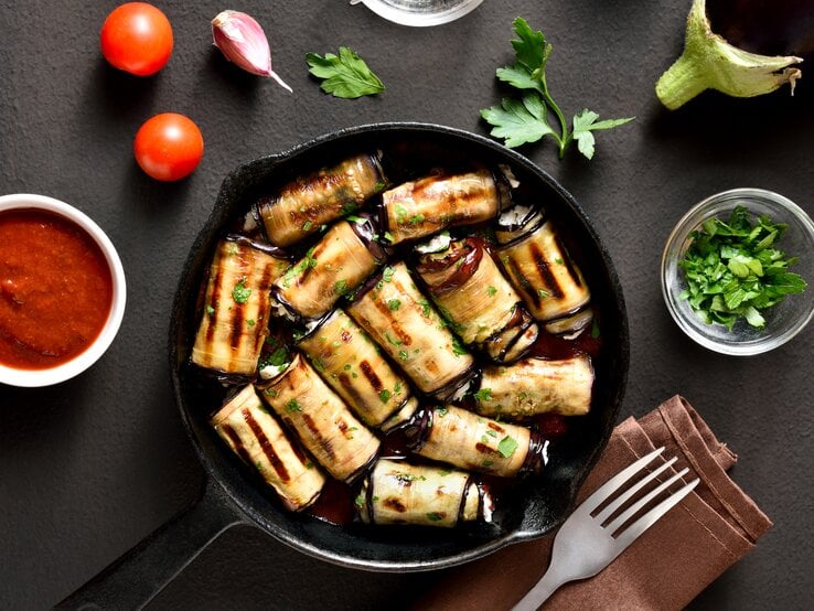 Gebratene Auberginen-Frischkäse-Röllchen in einer Pfanne. Drum herum liegen Tomaten, Petersilie, Knoblauchzehen sowie ein Dip und eine Gabel auf einem Tuch.