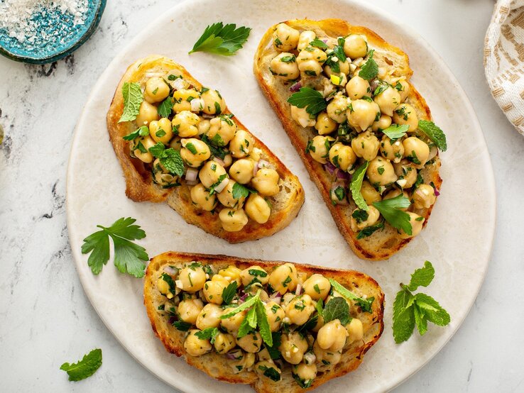 Ein weißer Teller mit drei Scheiben Bruschetta auf einem Marmoruntergrund direkt von oben fotografiert. Daneben ein weiß-braunes Tuch und etwas Salz und Olivenöl. | © Shutterstock/ Elena Veselova