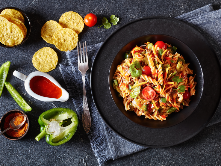 Nudelsalat mit Hackfleisch und Tacos auf schwarzem Teller, angerichtet auf dunklem Untergrund. Daneben liegen verschiedene Zutaten.  | © Getty Images/ from_my_point_of_view