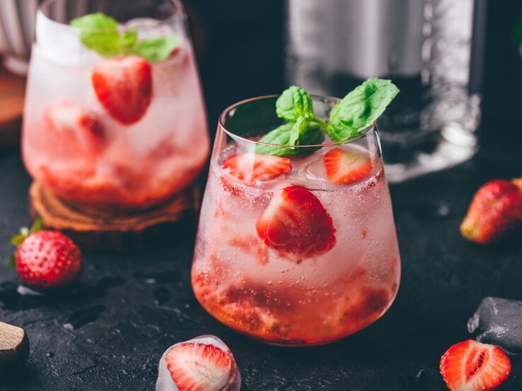 Erdbeer-Gin-Tonic auf einem dunklen Untergrund. Daneben liegen verschiedene Zutaten.