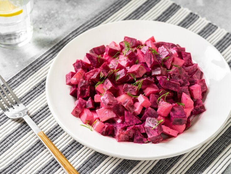 Ein Teller schwedischer Rote-Bete-Salat auf einem Platzdeckchen, daneben eine Gabel und ein Glas.
