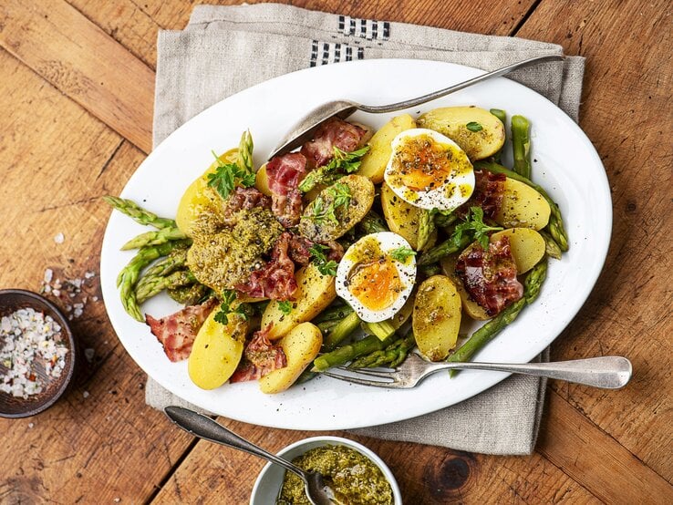 Kartoffelsalat mit gekochten Eiern, Pesto und knusprigem Pancetta und grünem Spargel auf einem weißen Teller mit Besteck. Daneben eine Schale mit Salz und mit Pesto. 