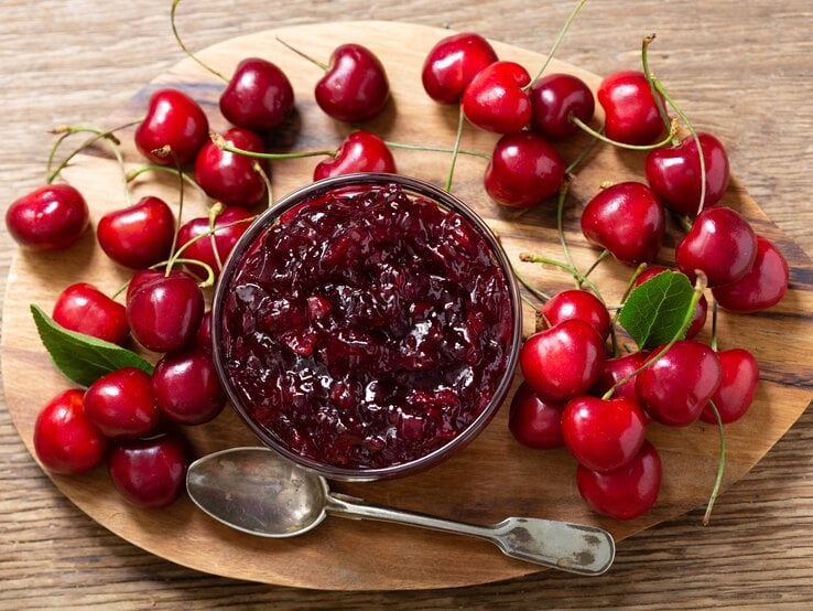 Auf einem Holzbrett steht ein Glas Kirschmarmelade. Drum herum liegen Kirschen.