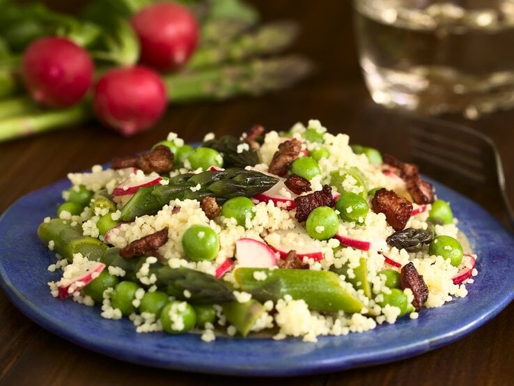 Eine Schale Spargel-Couscous-Salat mit Radieschen und Erbsen und Rosinen, drumherum die frischen Zutaten.