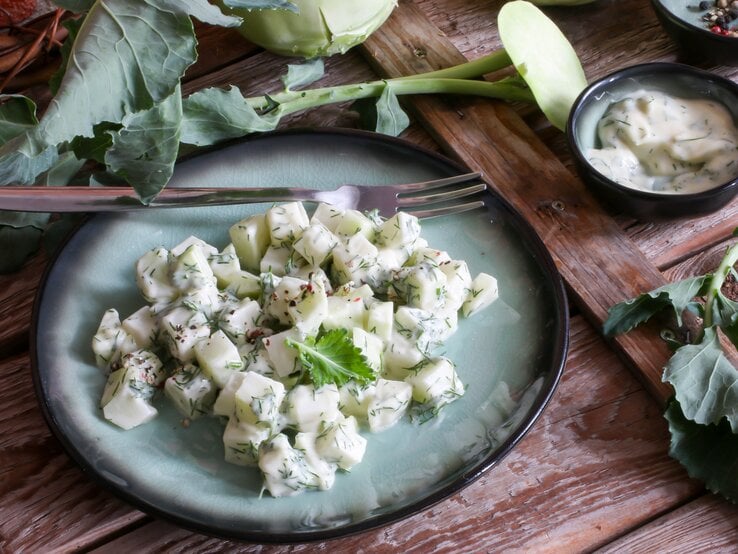 Schräge Draufsicht: ein blau-schwarzer Teller Kohlrabi-Joghurt-Salat steht auf einem dunklen Holztisch. Drum herum liegt frischer Kohlrabi. Und danaben ist ein Schälchen mit einer cremigen Soße.