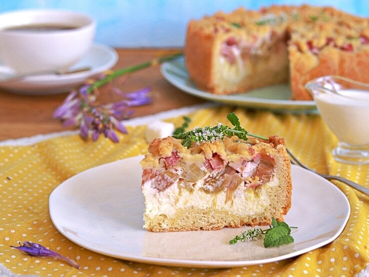 Ein Stück vom Rhabarberkuchen auf einem weißen Teller und einer gelb-blauen Tischdecke. Dahinter eine Tasse Kaffee und der angeschnittene Kuchen.  | © Shutterstock/ Happy_food_photo