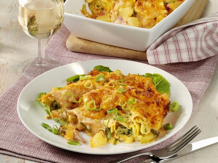 Auf einem Teller liegt eine Portion Tortellini-Auflauf 'Hawaii' mit Ananas, Schinken und Käse. Daneben steht ein Glas Weißwein.