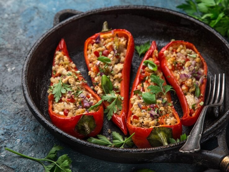 Eine gusseiserne Pfanne mit den vier gefüllten udn geschmorten Paprikaschoten auf einem dunkelgrauen Tisch mit Petersilie und einem grauen Tuch.