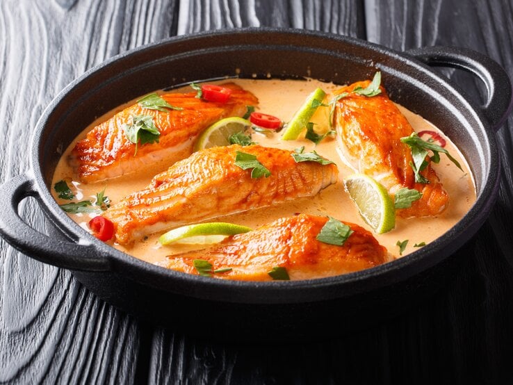 Eine schwarze Pfanne mit rotem Curry und Lachsfilet in Soße auf einem schwarzen Tisch.