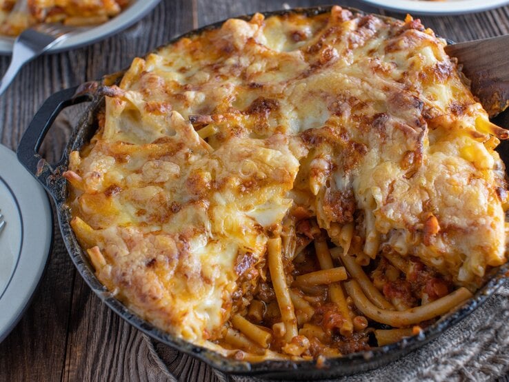 Makkaroni-Auflauf mit Hackfleischsoße und gestocktem Ei in schwarzer Auflaufform auf Holztisch. Daneben Teller mit Besteck und Auflauf. Draufsicht.
