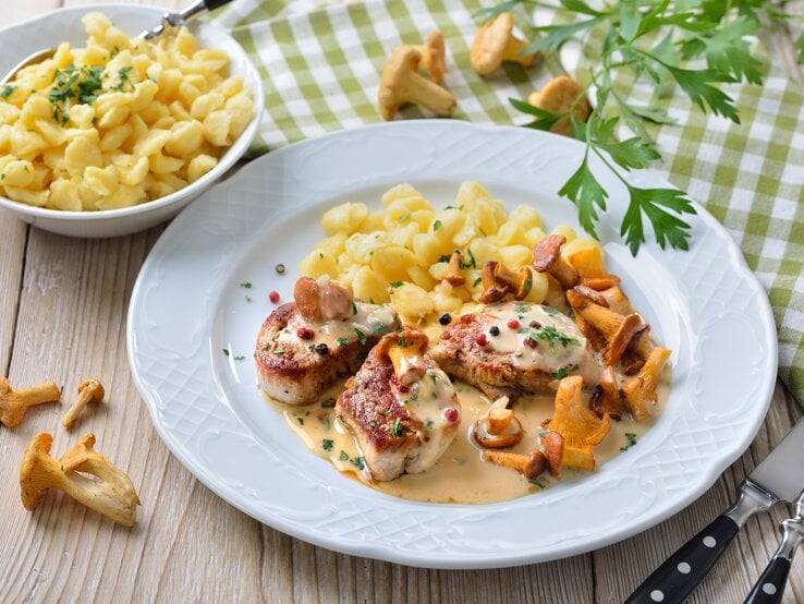Schweinemedaillons mit Pfifferlingen in Rahmsoße mit buntem Pfeffer und Knöpfle auf weißem Teller und Holztisch. Daneben Schale mit Knöpfle. Petersilie und Pfifferlinge als Dekoration auf dem Tisch verteilt. Aufnahme aus der Vogelperspektive. 