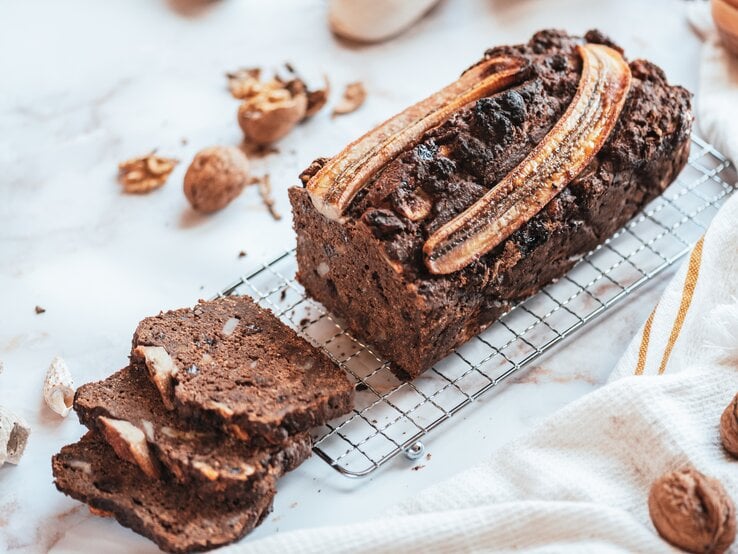 Ein angeschnittenes Schoko-Bananenbrot auf einem Kuchengitter, drumherum liegen geknackte Walnüsse.