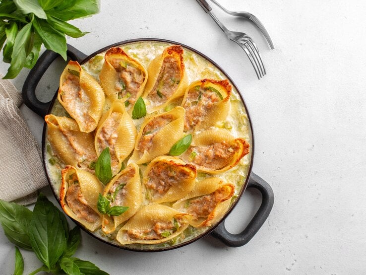 Draufsicht: Eine Auflaufform mit gefüllten Muschelnudeln mit Lachs und Schmand steht auf einem hellen Untergrund. Daneben liegen zwei gabeln, ein Glas mit frischem Basilikum und ein Leinentuch.