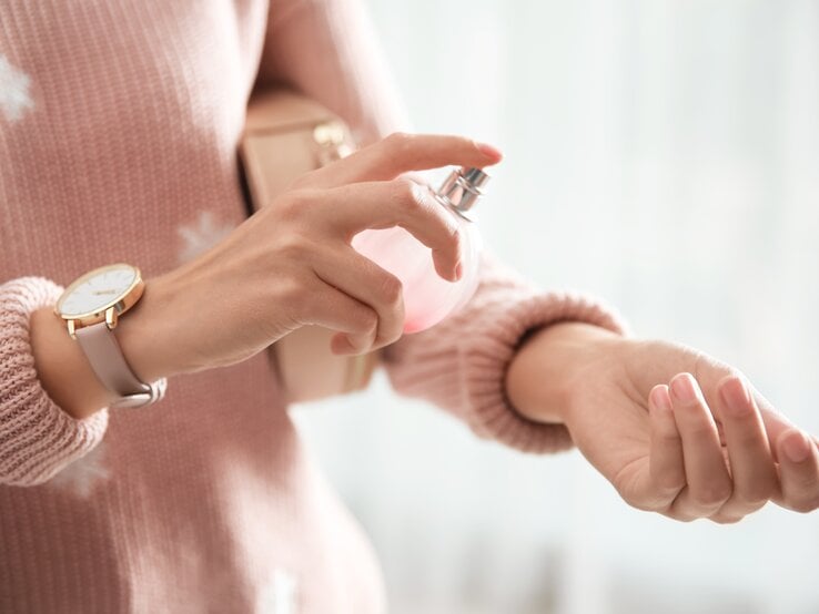 Eine Frau mit rosa Pullover sprüht Parfüm auf ihr Handgelenk. Das Parfüm steht im Fokus.