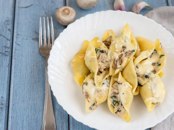 Draufsicht: auf einem hellen, sommerlich wirkenden Untergrund steht ein weißer Teller mit gefüllten Muschelnudeln mit Pilzen und Schmand. Daneben liegen eine Gabel und einige Pilze.