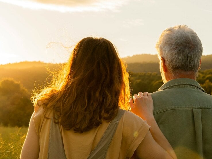 Zwei Personen von hinten, die gemeinsam in die Ferne blicken. Die Szene ist bei Sonnenuntergang oder Sonnenaufgang aufgenommen. Die Person auf der linken Seite hat schulterlanges, braunes Haar, während die Person auf der rechten Seite graues Haar hat. Die Person auf der linken Seite legt eine Hand auf die Schulter der anderen Person.