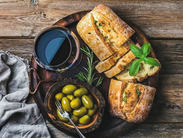Schnelles Olivenbrot auf dunklem Holzteller mit Glas Rotwein und Kräutern und Oliven auf Holztisch. Daneben graue Stoffserviette. Aufnahme aus der Vogelperspektive.