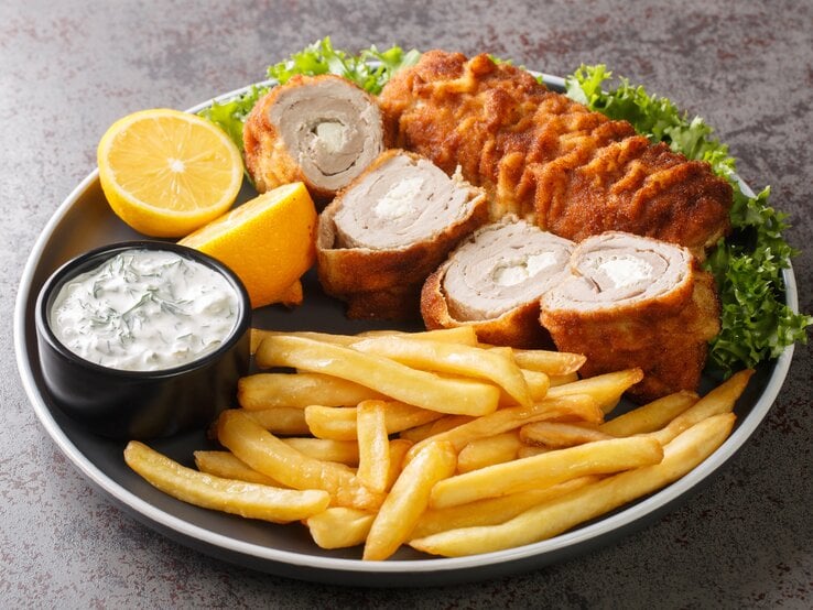 Serbisches Schnitzel mit Frischkäse-Füllung, dazu Pommes und Tatarsoße auf schwarzem Teller. 