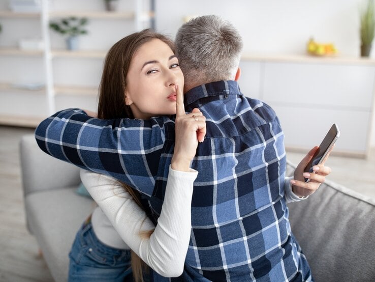 Frau und Mann umarmen sich und die Frau hat hinter dem Rücken des Mannes ein Handy in der Hand, guckt in die Kamera und macht das Hush-Zeichen.
