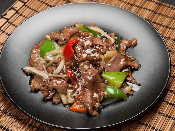 Ein schwarzer Teller mit dem Pfannengericht (Rindfleisch, Paprika, braune Soße) auf einer Bambusmatte und schwarzem Untergrund. Alles von schräg oben fotografiert. | © Shutterstock/ David Buzzard