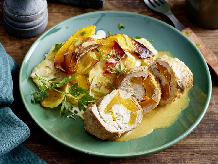 Schnitzelröllchen mit Kürbisgratin auf einem grauen Teller; auf dem Holzuntergrund ist eine Gabel, ein Tuch und eine Gewürzmühle.  | © EAT CLUB / Isabell Triemer 