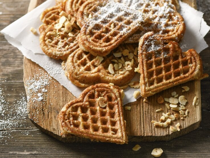 Waffeln in Herzform auf Krepp und hellbrauner Holzplatte angerichtet in der Draufsicht. Haselnusssplitter am Brettrand und den Waffeln verteil. Puderzucker darüber gestreut. Alles steht auf einem Holztisch mit dunkler Maserung. 