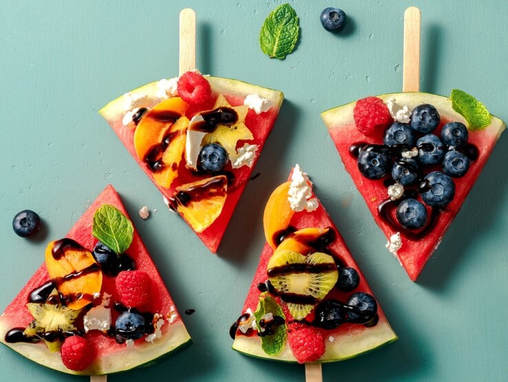 Vier Stücken Wassermelone, belegt mit Obst auf einem grünem Tisch. Alles direkt von oben fotografiert.