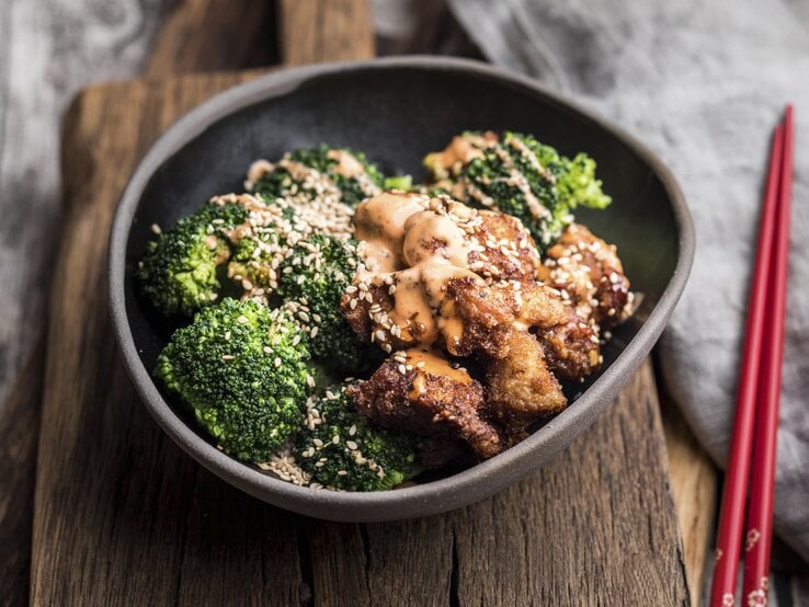 Schwarze Schüssel mit Brokkoli-Hähnchen-Pfanne mit Honig auf Holzbrett, daneben Stäbchen. | © Getty Images/ Westend61