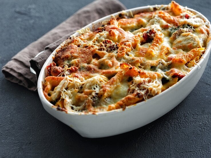 Kürbis-Nudelauflauf mit Spinat in einer ovalen Auflaufform, mit Käse überbacken, daneben ein graues Geschirrtuch.