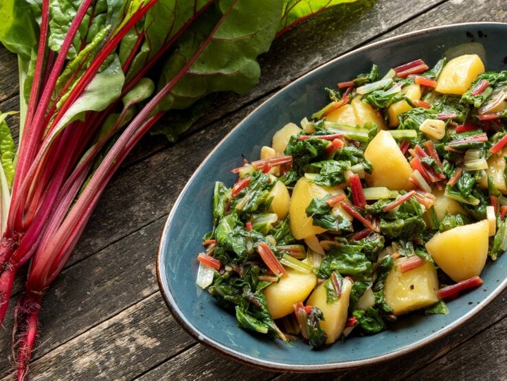 Ein blauer ovaler Teller mit der Kartoffel-Mangold-Pfanne auf dunklem Holztisch. Alles direkt von oben fotografiert.