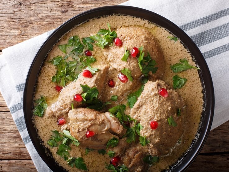 Ein Topf mit Satsivi, georgisches Gericht mit Hähnchen in Walnusssoße, garniert mit Petersilie und Granatapfelkernen, auf einem Holztisch, von oben. 