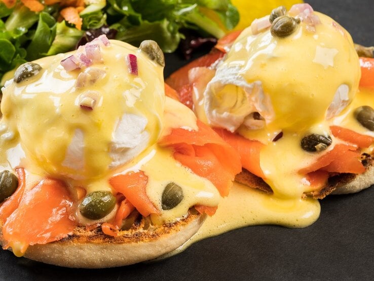 Die West-Coast-Eggs-Benedict auf schwarzem Untergrund mit einem kleinen Salat im Hintergrund. Alles seitlich und von leicht oben fotografiert.
