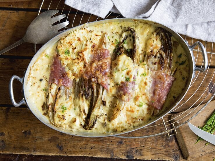 Keto Gratin mit Chicorée in Metall-Auflaufform auf Gitter, Holzuntergrund und Servierbesteck daneben. 