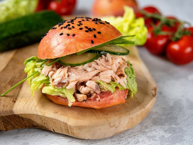 Pulled-Lachs-Burger auf einem Holzbrett. Im Hintergrund eine Gurke, Salat, Tomaten und Zwiebeln. 