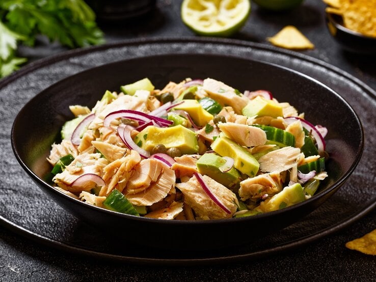 Ein schwarzer Teller mit dem Thunfischsalat, Avocado und Kapern auf einen schwarzen Tisch mit Limette, Nachos und  Petersilie.