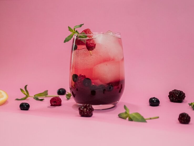 Ein Glas mit Lillet Spritz, Eiswürfeln, Beeren und Minze vor rosa Hintergrund, drumherum liegen Beeren und Minze.