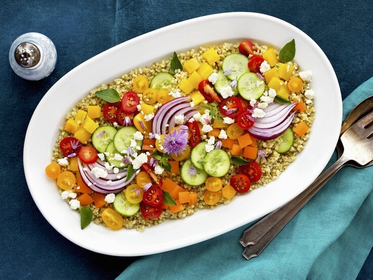 Bunter Salat aus Tomate, Paprika und Gurke auf Quinoa mit Fetakäse angerichtet auf einem weißen ovalen Porzellanteller, der auf blauer Tischedecke steht, neben dem Teller liegt Silberbesteck und steht ein Salzstreuer