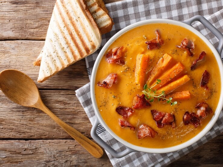 In einer großen Schüssel ist eine Süßkartoffel-Rahmsuppe. Die Suppenschüssel steht auf einem karierten Handtuch, dass auf einem Holztisch liegt. Daneben liegen zwei Scheiben Toast und ein Holzlöffel.