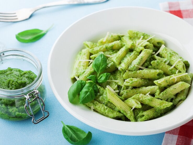 Ein Teller Penne mit grünem Miso-Pesto, Basilikum und Streukäse, daneben ein Glas mit dem Pesto sowie eine Gabel und eine rot-weiß-karierte Serviette.