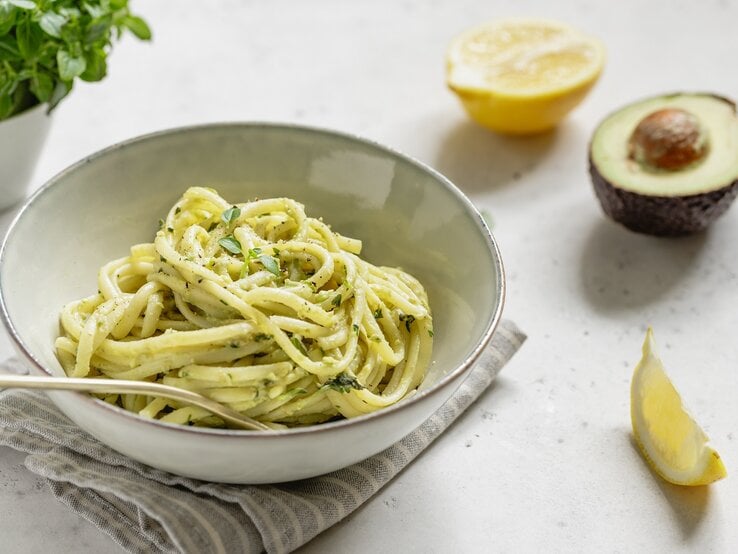Ein weißer Teller mit Avocado-Lemon-Pasta auf einem weißen Tisch neben frischer Avocado und Zitrone.