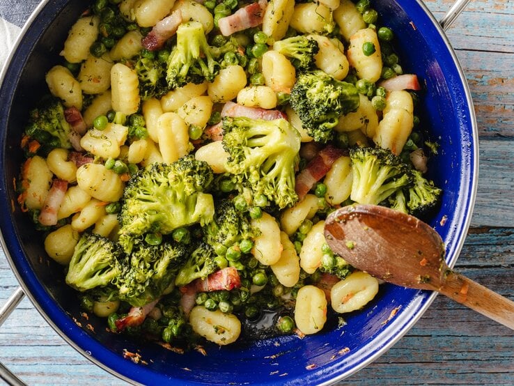One Pot Gnocchis in blauem Topf auf verblichen blau gestrichenem Holztisch. Daneben blau-weiß gestreiftes Leinentuch. Recht am Topf leicht darüber ragend ein Holzkochlöffel. Aufnahme aus der Vogelperspektive.