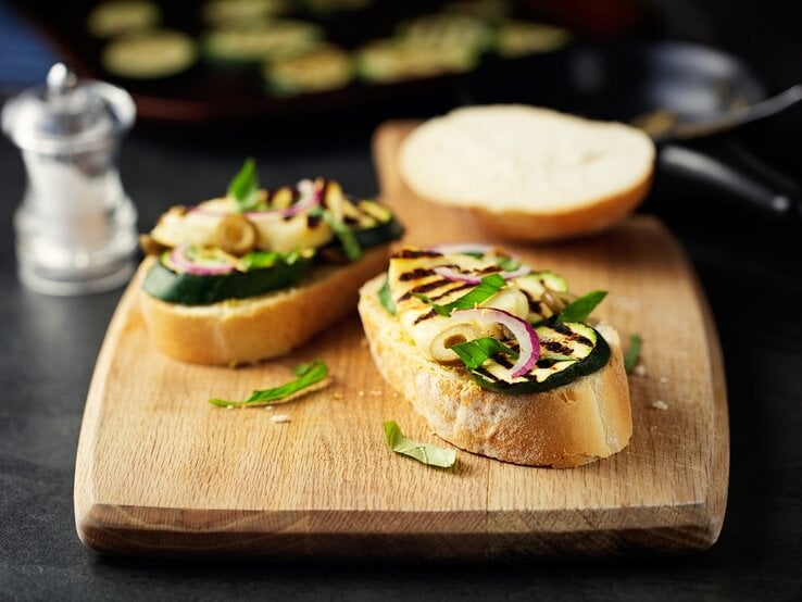 Ein Holzbrett mit Halloumi-Bruschetta mit Zucchini liegt auf einem dunklen Untergrund. Daneben steht ein Salzstreuer. | © Getty Images / haoliang
