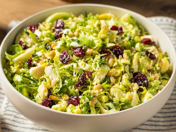 Eine weiße Schale mit dem Rosenkohl-Salat mit Cranberries und Pinienkernen auf einem blau-weiß gestreiftem Tuch und einem Holztisch von oben fotografiert.