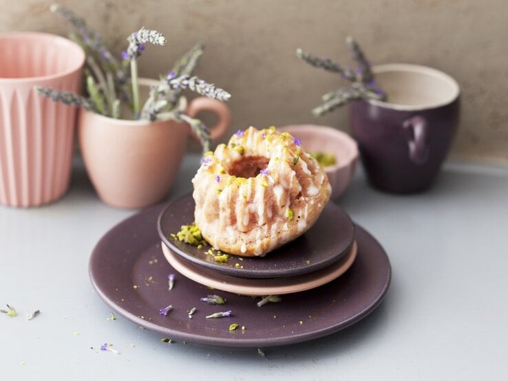 Ein Himbeer-Lavendel-Minigugelhupf auf Tellerstapel mit Geschirr und Lavendel im Hintergrund. Weißer Untergrund und beige Wand.
